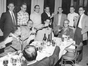 Men in suits and casual wear toast and smile around a dining table set with food, wine bottles, and glasses in a joyful scene.