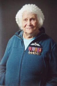 An elderly woman with white hair smiles gently, in a blue jacket with military medals and badges, against a dark background.