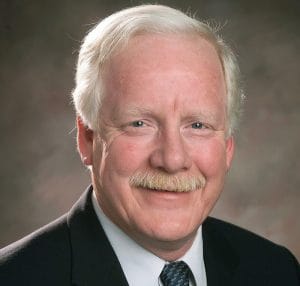 Portrait of an older man with short white hair, a light mustache, and fair skin, wearing a dark suit, white shirt, and patterned tie, smiling in front of a neutral studio background.
