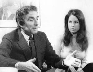 A man in a suit gestures as he speaks, seated beside a young woman with long hair. They are indoors by a plain wall and framed picture.