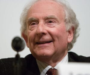 An older man with white hair smiles while seated at a microphone, wearing a dark jacket and tie. The plain background is blurred.