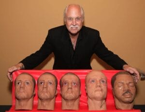 An older man with white hair and a mustache stands behind a table showing five bald, human-like head models on a red surface.