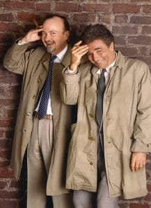 Two men in beige trench coats and ties stand before a brick wall, smiling and playfully touching foreheads; one holds a cigar.