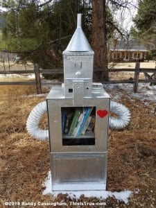 A metallic Little Free Library shaped like the Tin Man from “The Wizard of Oz,” with a heart symbol on its chest and flexible tube arms, stands outdoors by a fence and trees, holding books inside its chest compartment.
