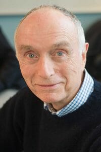 An older man with short gray hair, blue eyes, and light skin smiles slightly at the camera in a blue checkered shirt and dark sweater.