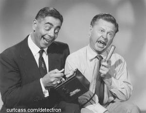 Two men in suits make exaggerated, silly faces as one holds an instrument's probe and the other operates the small electronic device. The scene is playful and comedic, in black and white.