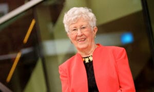 An older woman with short white hair and glasses smiles in a bright coral blazer over black, standing before a modern glass building.