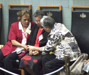 Three elderly women sit together on black chairs, holding hands and comforting each other. A roped barrier is visible in front.