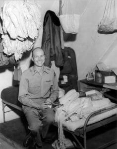 A man in military uniform smiles on a bed in a small room with parachutes, gear, and supplies scattered around him.