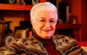 Older woman with short white hair and glasses smiles indoors, wearing a patterned sweater; books and decor fill shelves behind her.