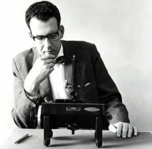 A man in glasses, a suit, and bow tie sits at a table, thoughtfully viewing a vintage spectrometer. A pen rests to his right.