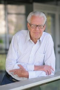 An older man with white hair and glasses, in a striped button-down, leans on an outdoor railing, calmly facing the camera.