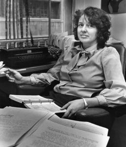 A woman with curly hair sits at a cluttered desk, holding a pen and open binder, smiling slightly; shelves and typewriter behind her.