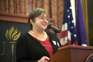 A woman with short gray hair and glasses speaks at a lectern with a microphone, wearing red, with U.S. flags and a sign reading YU.