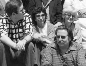 A black and white photo of four adults outdoors, sitting close, smiling and chatting. Three wear glasses; one has long hair and a mustache.