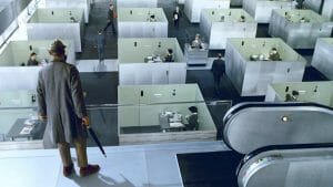 A man in a coat and hat stands atop an escalator, overlooking rows of gray office cubicles filled with people at work.