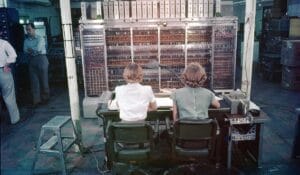 Two women sit at a control panel operating a large, 1950s computer with numerous wires and switches. Two men stand in the background amidst equipment, all in a room that exudes a historic, mid-20th-century ambiance.