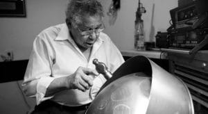 An elderly man in glasses and a white shirt shapes a steel drum with a hammer in a workshop, focused on his craft.