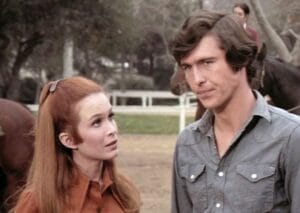 A woman with long red hair and a man with brown hair stand outdoors near horses, engaged in a serious conversation. In the background, there are trees and another person riding a horse.