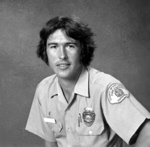 A young man with medium-length hair wears a light-colored uniform shirt adorned with a badge and an embroidered patch that reads "County of Los Angeles Fire Department." He sits in front of a plain studio backdrop.