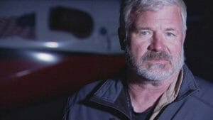 A middle-aged man with gray hair and a beard stands before a blurred aircraft, wearing a dark jacket and looking serious.