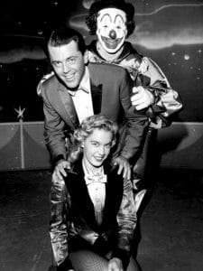 A man in a suit stands behind a seated woman in costume, both smiling, with a clown cheerfully posing behind them. The setting is a stage set.