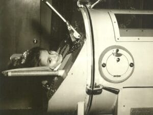 A young girl smiles with her head on a cushion, lying inside an iron lung that encases her body; only her head is visible.