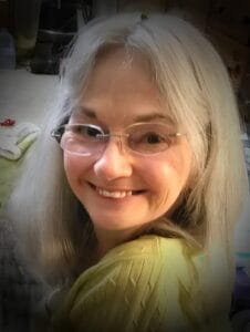 A woman with long gray hair and glasses smiles over her shoulder indoors, wearing a light green top in gentle natural lighting.
