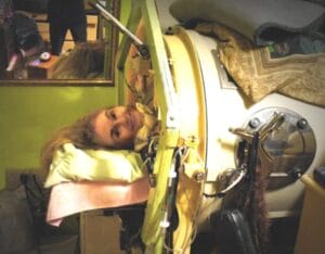 A person in an iron lung, head on pillows outside the device; green-walled room with a mirror and blankets atop the machine.