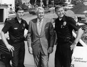 Three men pose in front of a police car; two actors in LAPD police uniforms and one in a suit. The setting appears to be outdoors on a sunny day, with a hill and buildings in the background.