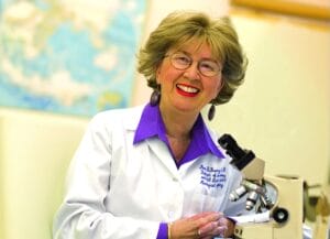 An older woman with glasses and short hair smiles in a white lab coat by a microscope in a lab, world map behind her.