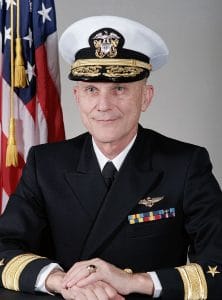 A U.S. Navy admiral in full dress uniform with medals, seated at a table, smiling, an American flag in the background.