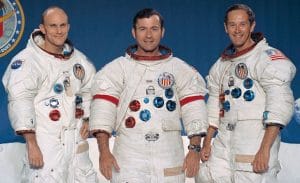 Three astronauts in white Apollo-era spacesuits stand side by side, smiling; their suits feature mission patches and flags, with a blue backdrop.