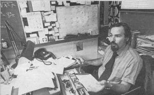 A man with a beard and mustache sits at a cluttered desk covered with papers and office supplies, surrounded by a bulletin board filled with notes and a large wall calendar. He appears to be speaking or explaining something.