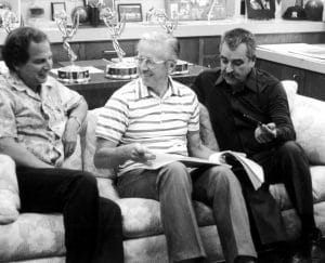Three men sit on a couch, smiling and talking, with one holding a book. Several Emmy awards are displayed on the cabinet behind them, along with framed photos and memorabilia.
