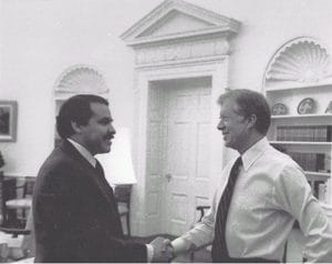 A man in business attire shake hands with President Jimmy Carter in the Oval Office of the White House, with bookshelves and decorative arches in the background.