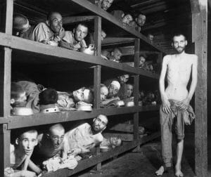 Emaciated men in striped uniforms lie crowded on wooden bunk beds in a concentration camp barrack; one man stands at the edge.