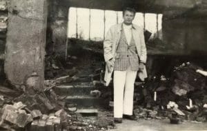 A person in a light coat and checked suit stands among rubble in a ruined building with broken windows, hinting at destruction.