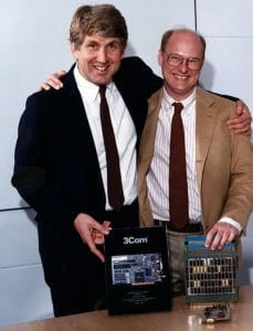 Two smiling men in suits stand together, one with an arm around the other, by a table displaying early 3Com computer hardware.