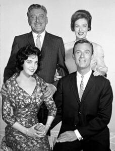Black-and-white photo of two men in suits and ties and two women in dresses, two standing behind and two seated, all smiling.