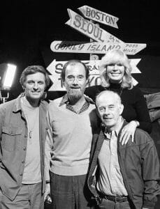 Four people smile together by a black-and-white signpost with arrows to cities like Boston, Seoul, and Coney Island on a TV set.