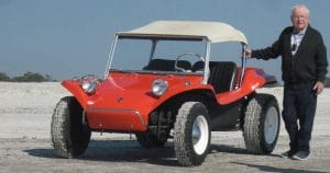 An elderly man stands by a classic red dune buggy with big tires and a white canopy, on sand beneath a clear blue sky.