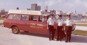 Three firefighters in uniform stand before a red City of Miami Rescue No. 2 vehicle, city skyline and water behind them.