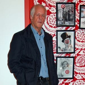 A gray-haired man in a dark jacket and blue shirt stands before a wall with red floral decor and celebrity photos.
