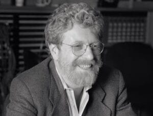 A man with curly hair, full beard, and glasses smiles indoors in a blazer and collared shirt, shelves and office gear behind him.