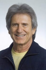 A smiling older man with graying hair, dressed in a dark jacket and yellow shirt, standing in front of a plain light background.