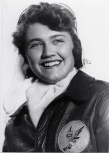A smiling woman wearing a leather flight jacket with a winged mascot patch, a scarf, and a flight cap with goggles, poses for a portrait.