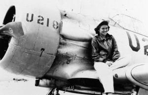 A woman in a leather jacket and white pants sits on the wing of a vintage military aircraft marked U218, smiling at the camera. The photo appears to be historical and in black and white.