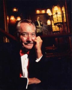 An older man with a mustache in a black tuxedo and red bow tie smiles, resting his head on his hand in an elegant, warm-lit room.