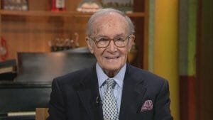 An elderly man with gray hair and glasses, in a dark suit and patterned tie, smiles seated in a warmly lit room with shelves and piano.